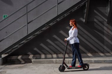 Beyaz gömlekli kızıl saçlı bir kız duvar boyunca elektrikli scooter kullanıyor. Pantolon kostümlü ve kırmızı topuklu ayakkabıgiyen bir iş kadını modern bir arabayla şehirde dolaşıyor. Scooter'da iş kadını.