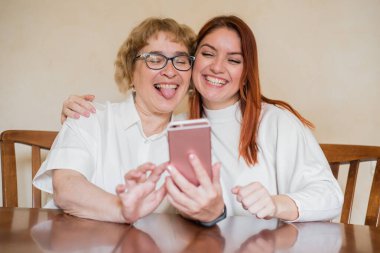 Anne ve kızı masada evde otururken akıllı telefonun ön kamera fotoğraflandı. Neşeli yaşlı anne ve kızı komik selfie'ler yapıyor ve dillerini gösteriyor.