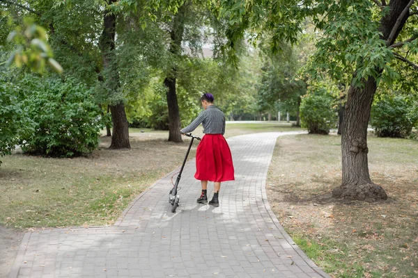一个紫色头发的年轻女子在公园里骑着电动滑板车。一个时髦的女孩穿着格子衬衫，一条红色的长裙和一条蝴蝶结，穿着一件现代装置，在城市中四处游荡。. — 图库照片