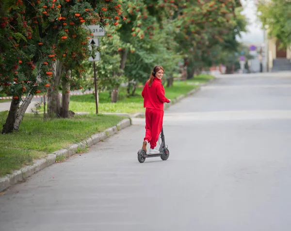 Chica pelirroja con un chándal rojo conduce un scooter eléctrico. Una joven con ropa de gran tamaño pasea por la ciudad y escucha música usando auriculares inalámbricos. Vista desde atrás . — Foto de Stock