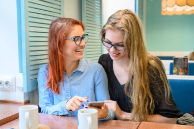 İki heyecanlı genç kız cep telefonu kullanıyor, bir kafede oturuyor ve parmakla işaret ediyor. Gözlüklü kızıl saçlı kadın akıllı telefon kız arkadaşının komik fotoğraflarını gösteriyor. Kahve içerler..