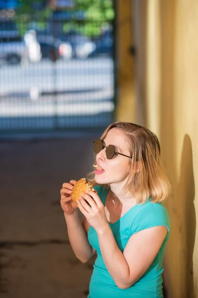 Portrét roztomilé dívky v zelených šatech venku a jíst Burger v teplém letním dnu. Krásná blondýnka v slunečních brýlích má na žluté stěně na ulici jídlo na smetí. Špatné stravní návyky. — Stock fotografie