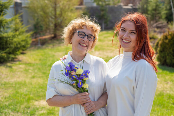 Взрослая дочь дарит цветы своей матери на улице, во дворе дома. Проводим время вместе, празднуем дома по выходным. День Матери. Теплые отношения между поколениями
