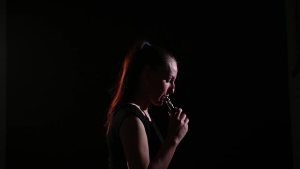 De brunette rookt een elektronische sigaret op een zwarte achtergrond, laat dikke rook uit haar mond ontsnappen. Portret van een vrouw die een vape rookt, zwevend. — Stockvideo