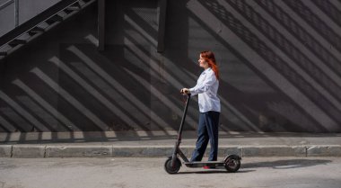 Beyaz gömlekli kızıl saçlı bir kız duvar boyunca elektrikli scooter kullanıyor. Pantolon kostümlü ve kırmızı topuklu ayakkabıgiyen bir iş kadını modern bir arabayla şehirde dolaşıyor. Scooter'da iş kadını.