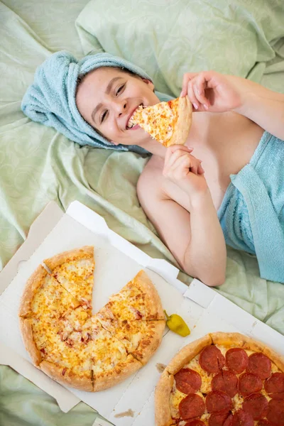 彼女の頭の上にタオルを持つかわいい女の子は、ベッドでピザを食べます。ベッドでピザを食べる若い女性。人生は喜び、体はポジティブです。イタリア料理が大好きです。食習慣、ファーストフード中毒. — ストック写真