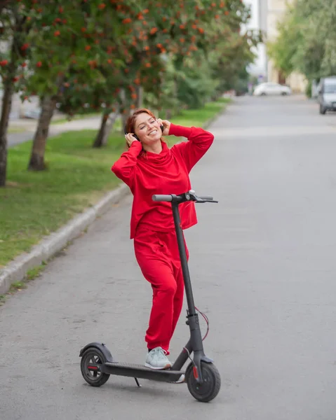 Рудоволоса дівчина в червоному костюмі керує електричним скутером. Молода жінка в надмірному одязі їде містом на сучасному транспорті і слухає музику за допомогою бездротових навушників . — стокове фото