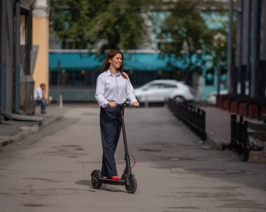 Beyaz gömlekli kızıl saçlı bir kız duvar boyunca elektrikli scooter kullanıyor. Pantolon kostümlü ve kırmızı topuklu ayakkabıgiyen bir iş kadını modern bir arabayla şehirde dolaşıyor. ofiste kıyafet kodu.