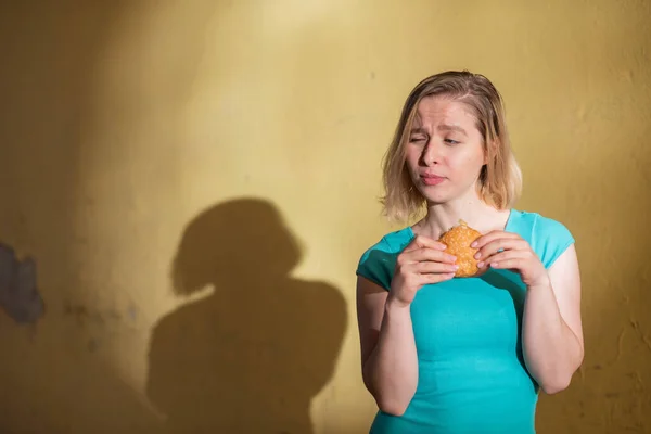 Porträtt av en söt flicka i en grön klänning gå utomhus och äta en hamburgare på en varm sommar dag. Vacker blondin njuter snabbmat på den gula väggen utanför. Fel matvanor. — Stockfoto