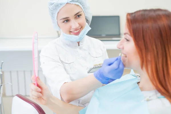 Verifique e selecione a cor dos dentes na cadeira do dentista. Dentista faz o processo de tratar uma bela jovem ruiva. Seleção de um implante dentário. Paciente olhando no espelho . — Fotografia de Stock