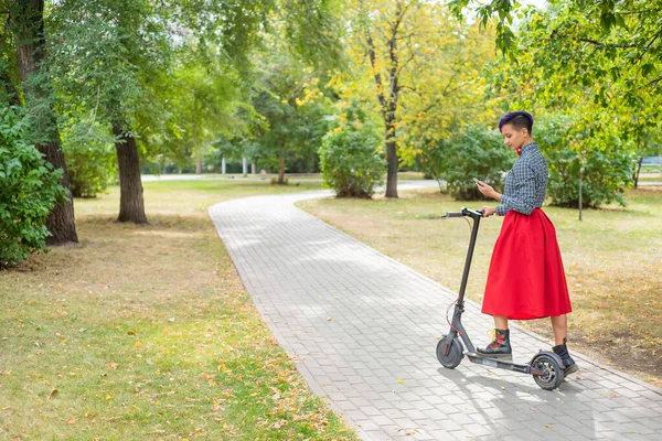 A fiatal nő, lila hajú lovaglás elektromos robogó a parkban. Stílusos lány egy borotvált templom egy kockás ing, egy hosszú vörös szoknya és csokornyakkendő írja egy üzenetet a smartphone. Csípő — Stock Fotó