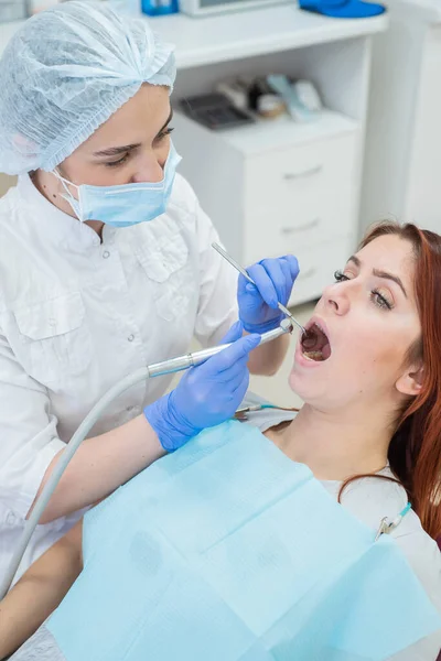 Una atractiva doctora sonriente en uniforme blanco examina a una paciente de cabello rojo. Dentista produce trata los dientes con un taladro . — Foto de Stock
