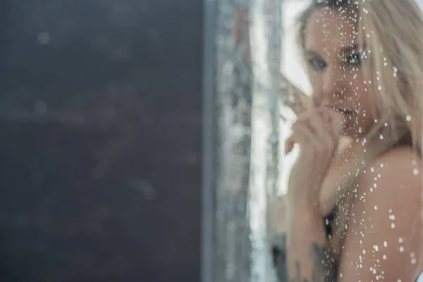 Europeo ragazza in biancheria intima nera, premuto il petto e le mani per il vetro bagnato e leccare erotico lingua gocce. — Foto Stock