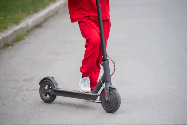 Рыжая девушка в красном спортивном костюме водит электрический скутер. Молодая женщина в огромной одежде разъезжает по городу на современном транспорте. Закрыть . — стоковое фото