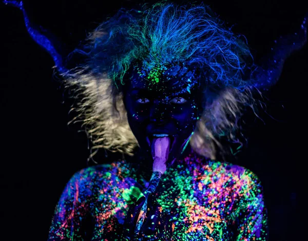 The girl in the image of the devil, contact lenses and horns. Woman paints her tongue in ultraviolet paint. fluorescent powder. Body art glowing in ultraviolet light. Stars in the eyes.