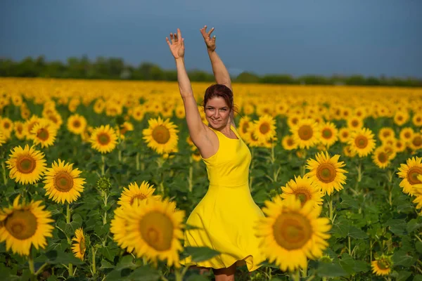 Rothaarige Frau in einem gelben Kleid, die mit erhobenen Händen in einem Sonnenblumenfeld tanzt. schöne Mädchen im Rock Sonne genießt einen wolkenlosen Tag auf dem Land. rosa Haarsträhnen. — Stockfoto