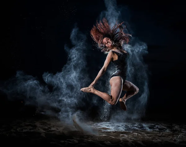 Danseuse de ballet en body noir saute haut et danse avec de la farine sur la plage le soir. Ballet de rue féminin. Farine, talc, poussière. Jeune femme rousse sautant dans un nuage de poudre bleue . — Photo