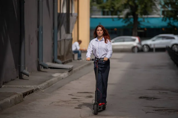 A vörös hajú lány egy fehér inget vezet elektromos robogó a fal mentén. Egy üzletasszony egy nadrág öltöny és piros magas sarkú túrák körül a várost a modern autó. öltözködési az irodában. — Stock Fotó