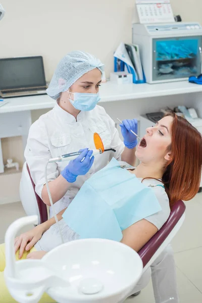 Dentista femenina fija y seca los rellenos dentales con luz. Hermosa dentista femenina poniendo un relleno en un diente con caries de una paciente de cabello rojo —  Fotos de Stock