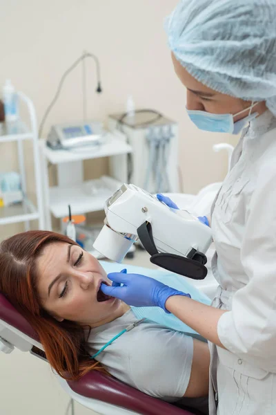 Atractiva doctora sonriente en uniforme blanco con herramientas en la mano. Joven dentista hermosa toma rayos X de mandíbula de paciente femenina. Mujer atractiva paciente en el dentista que toma examen de rayos X . —  Fotos de Stock