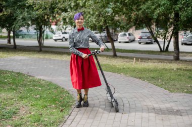 Mor saçlı genç bir kadın parkta elektrikli scooter kullanıyor. Ekose gömlek, uzun kırmızı etek ve papyon şık kız modern bir cihaz üzerinde şehir etrafında sürme. Hipster.
