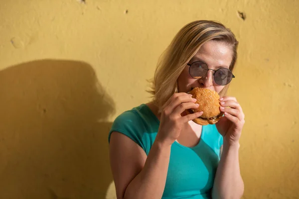 Yeşil bir elbise dışarı yürüyüş ve sıcak bir yaz gününde bir burger yeme sevimli bir kız portresi. Güneş gözlüğü güzel sarışın sokakta sarı duvar önemsiz yiyecek sahiptir. Yanlış gıda alışkanlıkları. — Stok fotoğraf