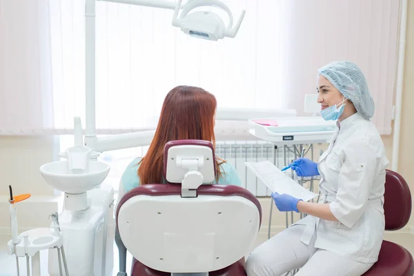 Joven dentista hermosa hablando con una paciente en el consultorio. Una mujer pelirroja se sienta en una silla con un ortodoncista y escucha las citas. Servicios dentales. Vista desde atrás —  Fotos de Stock