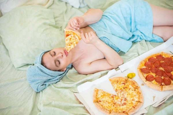 Menina bonito com uma toalha na cabeça come pizza na cama. Jovem mulher a comer pizza na cama. A vida é um prazer, corpo positivo. Amor pela comida italiana. Hábitos alimentares, dependência de fast food . — Fotografia de Stock