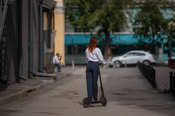 Rudowłosy dziewczyna w białej koszulce napędza skuter elektryczny wzdłuż ściany. Kobieta biznesu w garniturze spodni i czerwone wysokie obcasy jeździ po mieście w nowoczesnym samochodzie. kod stroju w biurze. — Zdjęcie stockowe