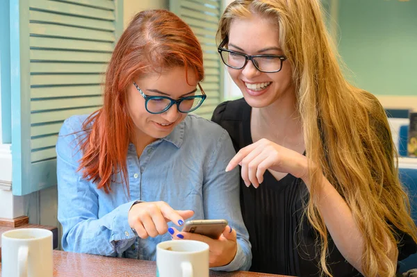 İki heyecanlı genç kız cep telefonu kullanıyor, bir kafede oturuyor ve parmakla işaret ediyor. Gözlüklü kızıl saçlı kadın akıllı telefon kız arkadaşının komik fotoğraflarını gösteriyor. Kahve içerler.. — Stok fotoğraf