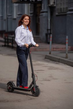 Beyaz gömlekli kızıl saçlı bir kız duvar boyunca elektrikli scooter kullanıyor. Pantolon kostümlü ve kırmızı topuklu ayakkabıgiyen bir iş kadını modern bir arabayla şehirde dolaşıyor. ofiste kıyafet kodu.