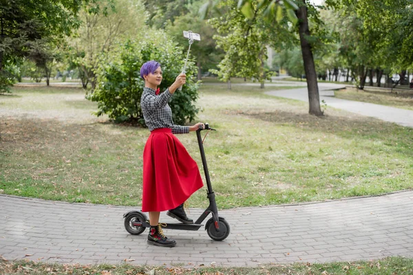 Una joven con el pelo morado monta una scooter eléctrica en un parque. Chica con estilo con un templo afeitado en una camisa a cuadros, una falda larga de color rojo y una corbata de lazo toma una foto con un palo de selfie. Hipster. . — Foto de Stock