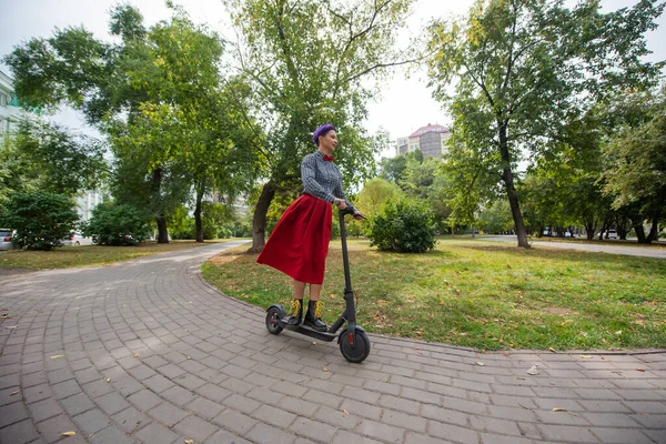 Egy fiatal nő, lila hajú lovagol egy elektromos robogó egy parkban. Egy stílusos lány egy borotvált templom egy kockás ing, egy hosszú vörös szoknya és egy csokornyakkendő a lovaglás a város körül egy modern eszköz. — Stock Fotó