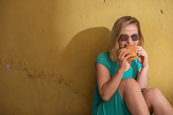 Šťastná žena v tyrkysově šatech sedí na chodníku a sní Burger v teplém letním dnu. Krásná blondýnka v slunečních brýlích má na žluté stěně obytné budovy podřadné pokrmy. — Stock fotografie