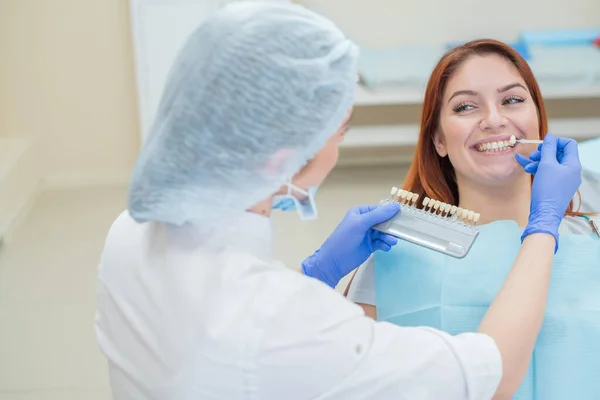 Verifique e selecione a cor dos dentes na cadeira do dentista. Dentista faz o processo de tratar uma bela jovem ruiva. Seleção de um implante dentário . — Fotografia de Stock