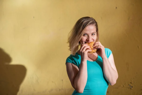 Retrato de una linda chica en un vestido verde caminando al aire libre y comiendo una hamburguesa en un cálido día de verano. Hermosa rubia está disfrutando de la comida rápida en la pared amarilla exterior. Hábitos alimenticios incorrectos . — Foto de Stock