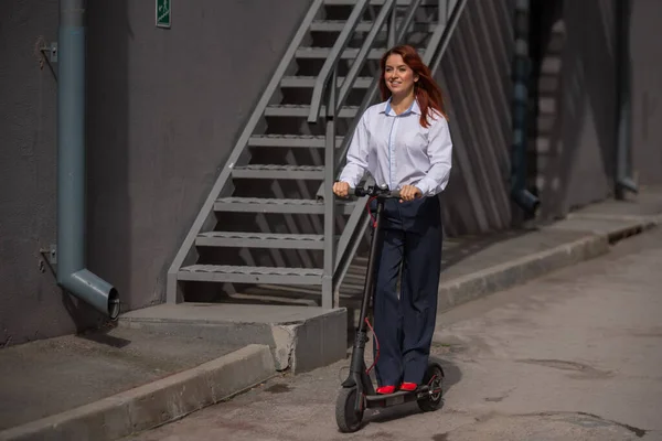 Una chica pelirroja con camisa blanca conduce un scooter eléctrico a lo largo de la pared. Una mujer de negocios con un traje de pantalón y tacones altos rojos pasea por la ciudad en un coche moderno. código de vestimenta en la oficina . —  Fotos de Stock