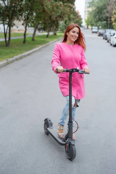 Yırtık kot pantolon ve yüksek topuklu sandalet kırmızı saçlı genç bir kadın yolda bir elektrikli scooter sürmek. Pembe ceketli bir kız hızlı bir şekilde uygun bir elektrikli ulaşım ile şehir etrafında hareket eder. — Stok fotoğraf