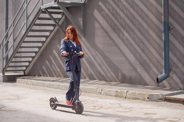 A vörös hajú lány egy kabátot és nadrágot vezet elektromos robogó a fal mentén. Egy üzletasszony egy pantsuit és piros magas sarkú túrák körül a várost a modern autó. Irodai dolgozó. Öltözködési kód — Stock Fotó