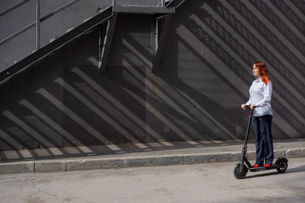Beyaz gömlekli kızıl saçlı bir kız duvar boyunca elektrikli scooter kullanıyor. Pantolon kostümlü ve kırmızı topuklu ayakkabıgiyen bir iş kadını modern bir arabayla şehirde dolaşıyor. Scooter'da iş kadını. — Stok fotoğraf