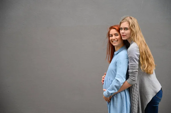 Vztahy stejného pohlaví. Červenovlasá těhotná žena v džínovových šatech stojí proti šedé zdi a její ruka jí odpočívá pod břicho. Žena se jemně objala matku zezadu. — Stock fotografie