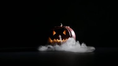 Cadılar Bayramı, koyu arkaplanlı korkutucu parlak yüzlü turuncu balkabağı. Kalın gri duman sarsıcı bir balkabağından çıkar. Tüm azizlerin arifesinde bir fenerin yakın çekimi