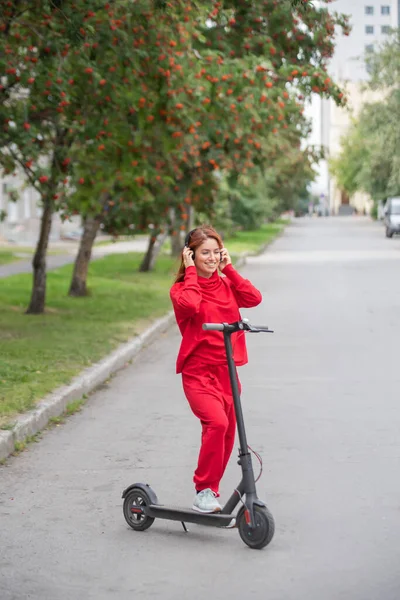 Chica pelirroja con un chándal rojo conduce un scooter eléctrico. Una joven en ropa de gran tamaño pasea por la ciudad en transporte moderno y escucha música usando auriculares inalámbricos . — Foto de Stock