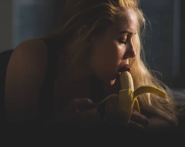 Das Konzept des Oralsex. Mädchen mit blonden Haaren, sexy Lecken und Liebkosen Lippen und Zunge große Banane auf dem Bett im Schlafzimmer. Erotische Fantasie. — Stockfoto