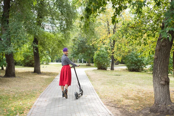 Een jonge vrouw met paars haar berijdt een elektrische scooter in een park. Stijlvol meisje in een geruite shirt, een lange rode rok en een strikje is rijden rond de stad op een modern apparaat. Hipster. — Stockfoto