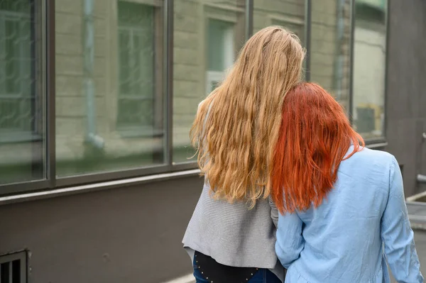 Två vänner går längs gatan och kramar varsamt varandra runt midjan. Blondinen i jackan och de rödhåriga damernas klänning böjde sina huvuden mot varandra. Utsikt från baksidan — Stockfoto