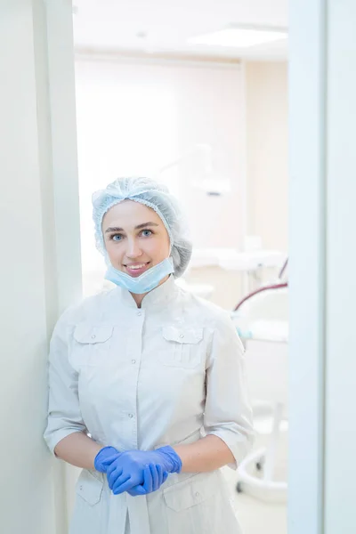 Attraente medico donna sorridente in uniforme bianca con i guanti è in piedi sulla porta dell'ufficio. dentista donna nel corridoio di una clinica con cappello e maschera . — Foto Stock