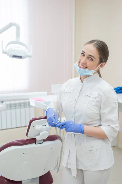 Ελκυστική, χαμογελαστή γυναίκα γιατρός με λευκή στολή στο χώρο εργασίας. Νεαρό όμορφο θηλυκό οδοντίατρο με μάσκα και γάντια στέκεται στο γραφείο κοντά στην πολυθρόνα με εργαλεία στα χέρια της — Φωτογραφία Αρχείου