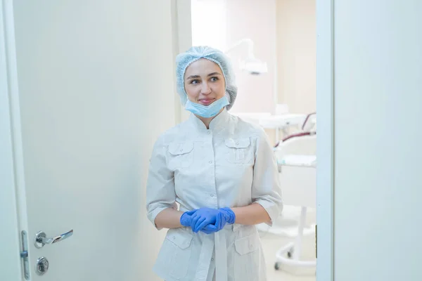 Attraktiv leende kvinnlig doktor i vit Uniform i handskar står i dörröppningen på kontoret. kvinna tandläkare i korridoren på en klinik i en mössa och mask. — Stockfoto