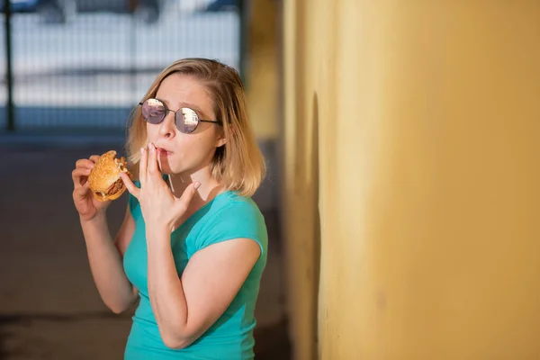 Portrét roztomilé dívky v zelených šatech venku a jíst Burger v teplém letním dnu. Krásná blondýnka v slunečních brýlích má na žluté stěně na ulici jídlo na smetí. Špatné stravní návyky. — Stock fotografie